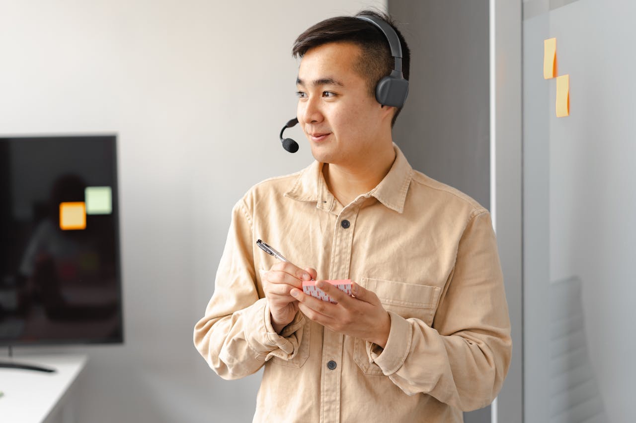 Smiling Asian man with headset holding notes, representing customer service in an office.